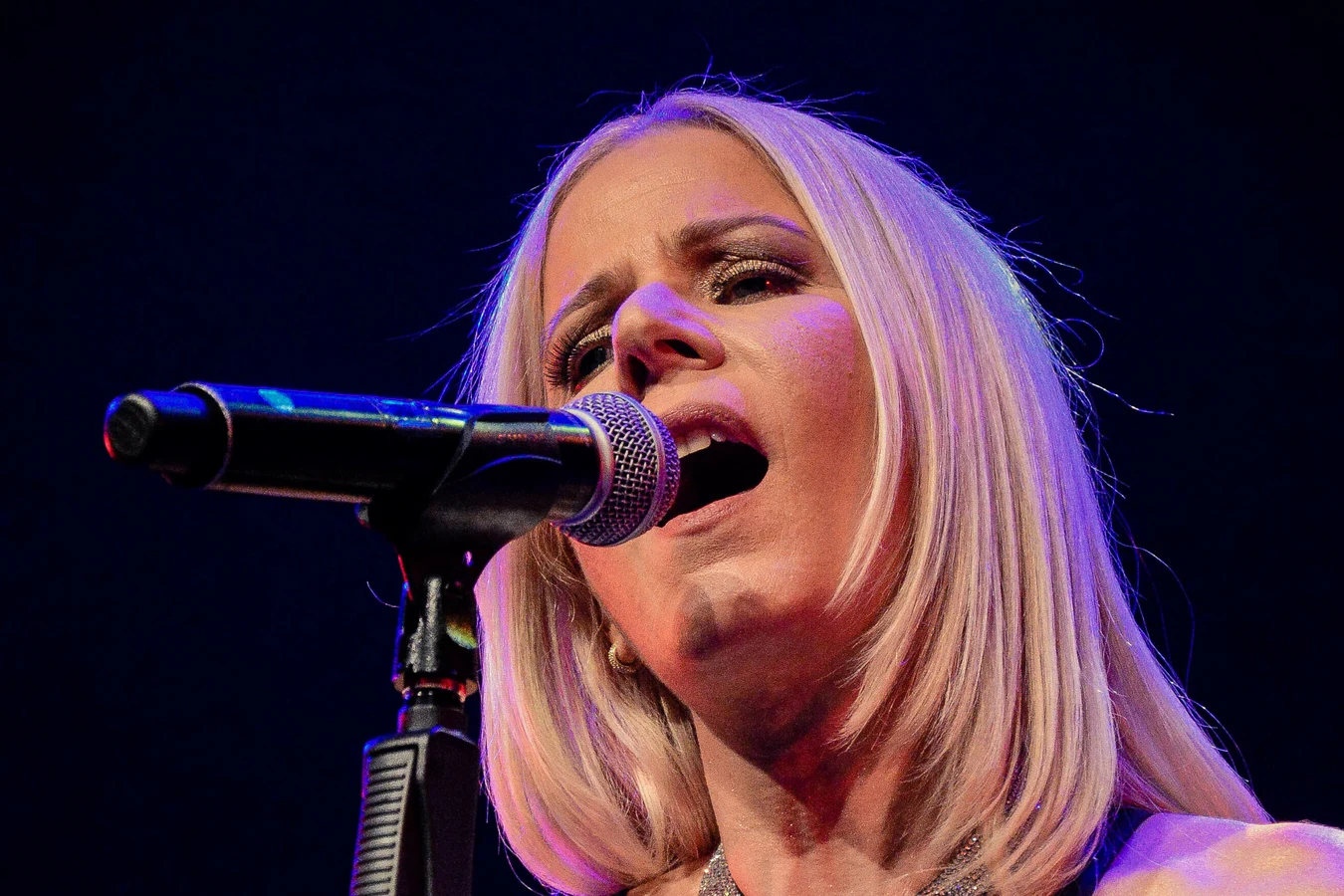 Paula Toller cantando ao microfone no palco, sob iluminação azul e violeta